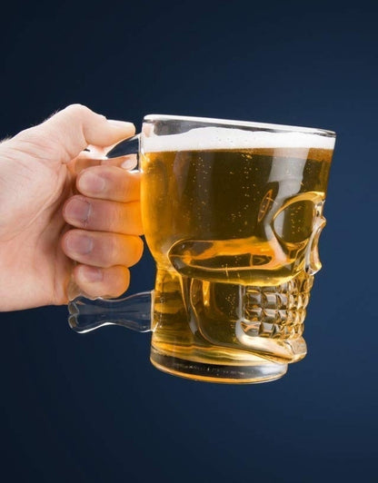 Hand holding a skull-shaped glass mug filled with beer against a dark background.
