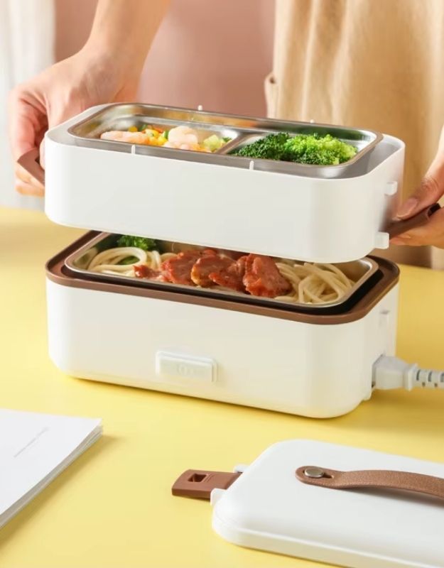 A person is opening a white portable electric heating lunch box with two tiers, showing the contents of the box which include food items like noodles and vegetables. The lunch box is designed for heating food without a microwave.