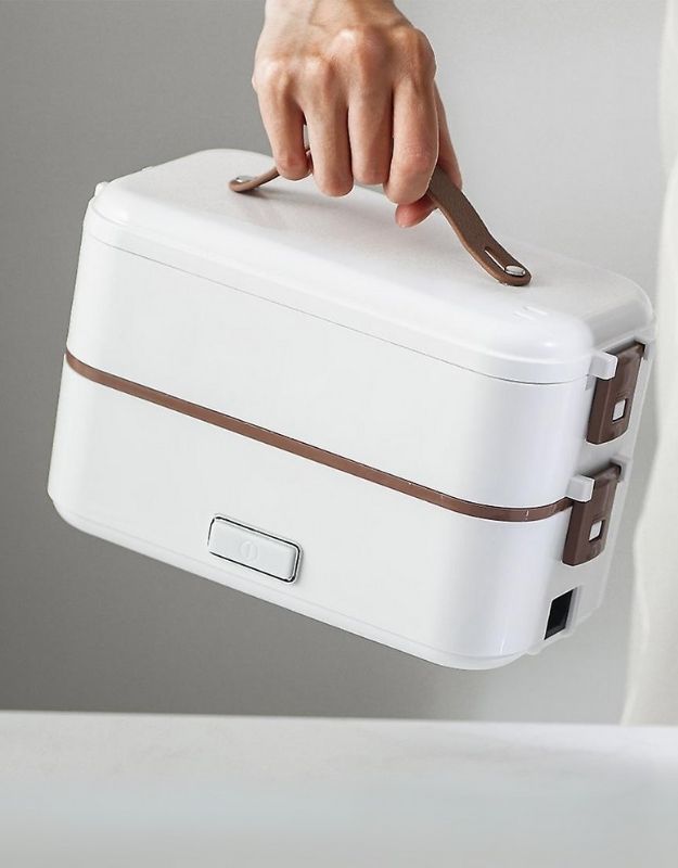 A person holding a modern, compact, white-colored lunch box with a brown strap-style handle. The lunch box has two tiers, held together with secure brown clasps on the side.