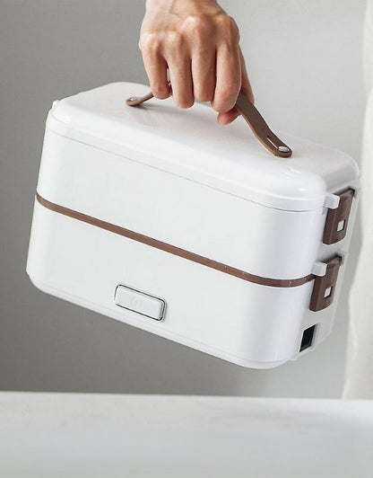 A person holding a modern, compact, white-colored lunch box with a brown strap-style handle. The lunch box has two tiers, held together with secure brown clasps on the side.