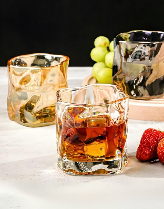 Glass of whiskey with ice cubes on a white surface with fruits in the background.