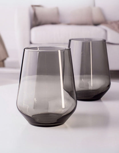 Two smoked glass tumblers on a white surface with a blurred background. Two smoky gray drinking glasses on a white table in a modern living room.