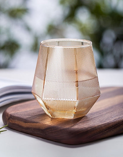 Gold mesh candle holder on a wooden cutting board with a blurred natural background. Amber faceted drinking glass with textured design on wooden board.