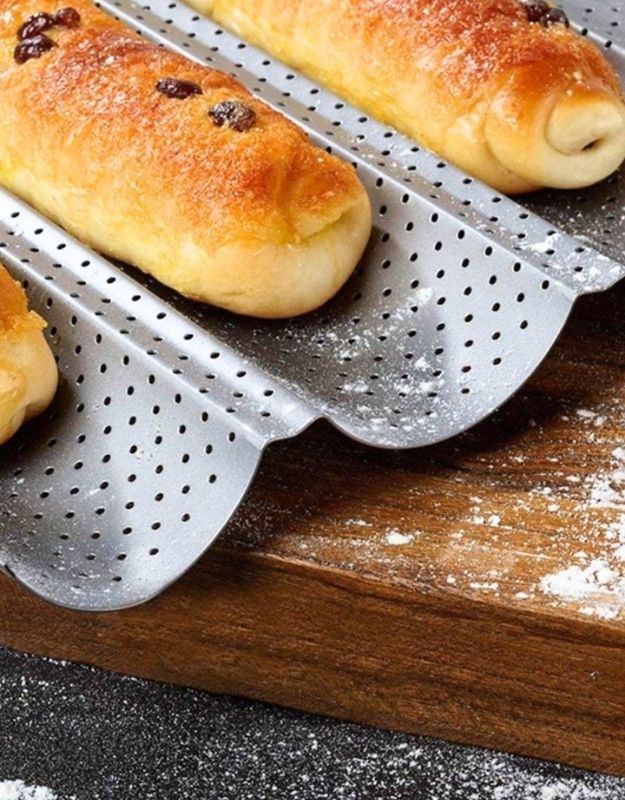 Baked pastries on a perforated baking sheet with a wooden surface underneath. Perforated baguette pan baking fresh bread rolls.