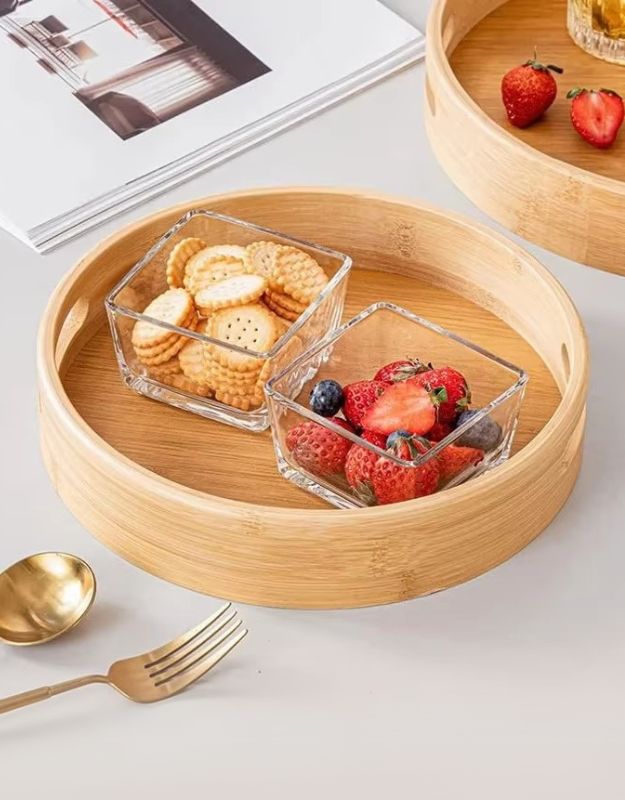 A round bamboo wooden serving tray with handles, featuring compartments with fruits and snacks on a table.
Round bamboo serving tray with glass bowls of cookies and fresh berries.