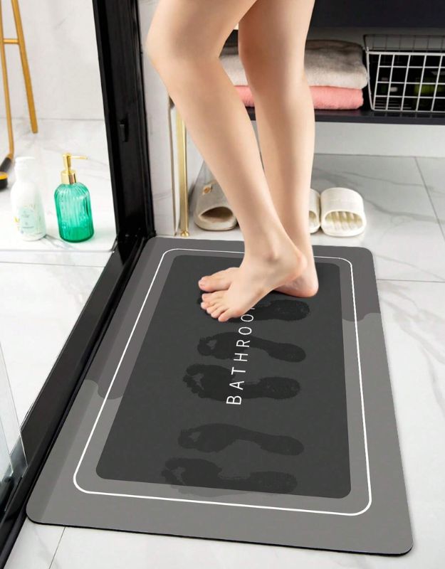 A person standing on a black bath mat with the text 'BATHROOM' written on it in white letters. The mat is placed on a tiled bathroom floor.