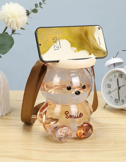Bear-shaped water bottle in use as a phone stand on a desk with flowers and a clock.
