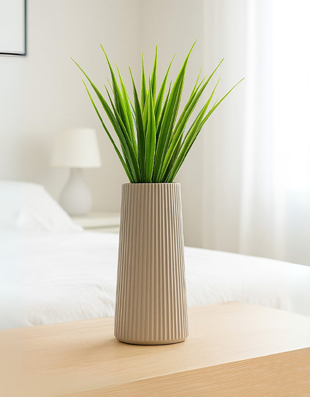 Neutral beige striped vase with green artificial reeds on a wooden surface in a softly lit modern bedroom.
