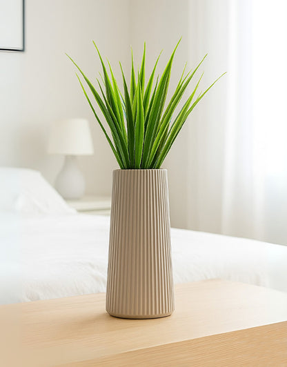 Neutral beige striped vase with green artificial reeds on a wooden surface in a softly lit modern bedroom.