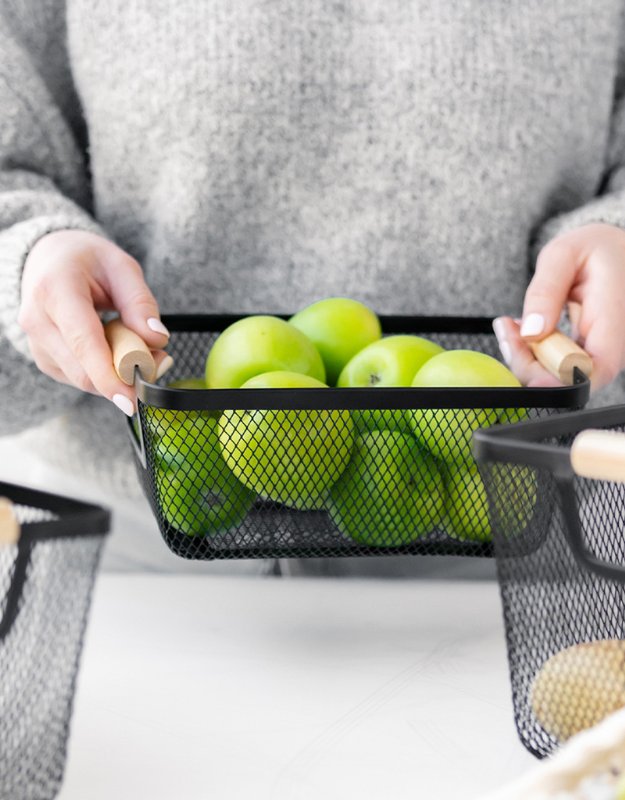 A rectangular metal mesh basket with a wooden handle filled with green apples, held by a person wearing a gray sweater.