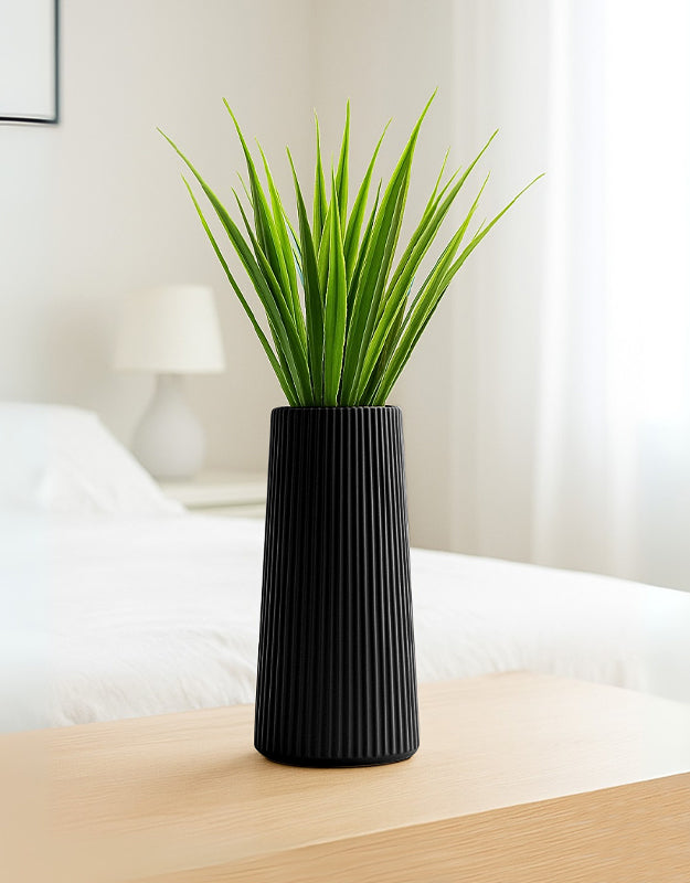 Minimalist black striped vase with green artificial reeds placed on a wooden table in a modern bedroom setting.