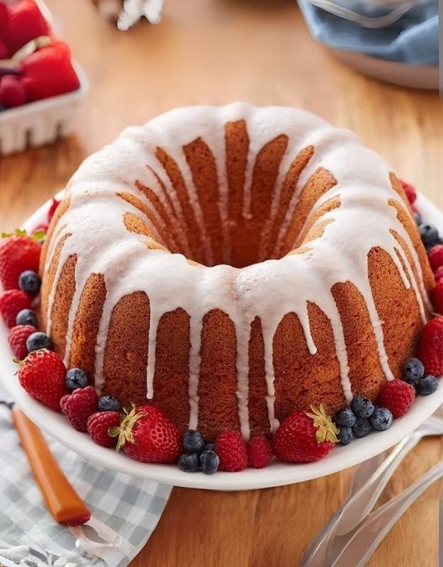 Bundt cake with white icing on a plate surrounded by berries on a wooden table. Bundt cake with icing and fresh berries baked in fluted red Bundt pan.