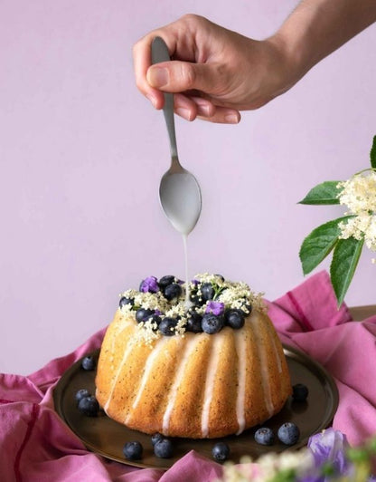 Bundt cake with blueberries being drizzled with a white sauce on a pink background. Freshly baked Bundt cake decorated with icing, blueberries, and edible flowers.