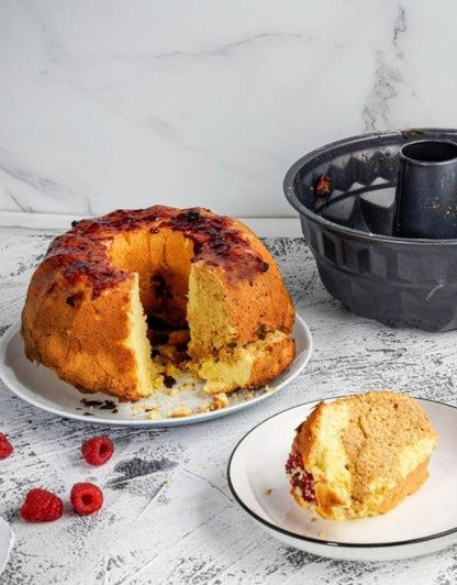 Baked cake with a slice taken out, placed on a white plate with raspberries, next to a black baking mold on a marble countertop. Freshly baked bundt cake next to nonstick bundt cake pan with tube design.
