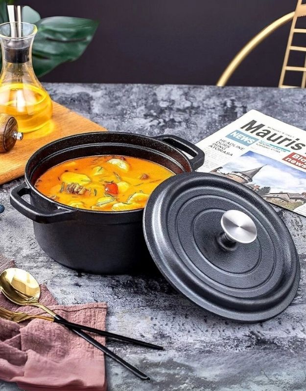 Black cast iron pot with a lid containing a hot dish on a gray surface with a magazine in the background. Black cast iron cooking pot with lid, filled with vegetable stew on a kitchen table.