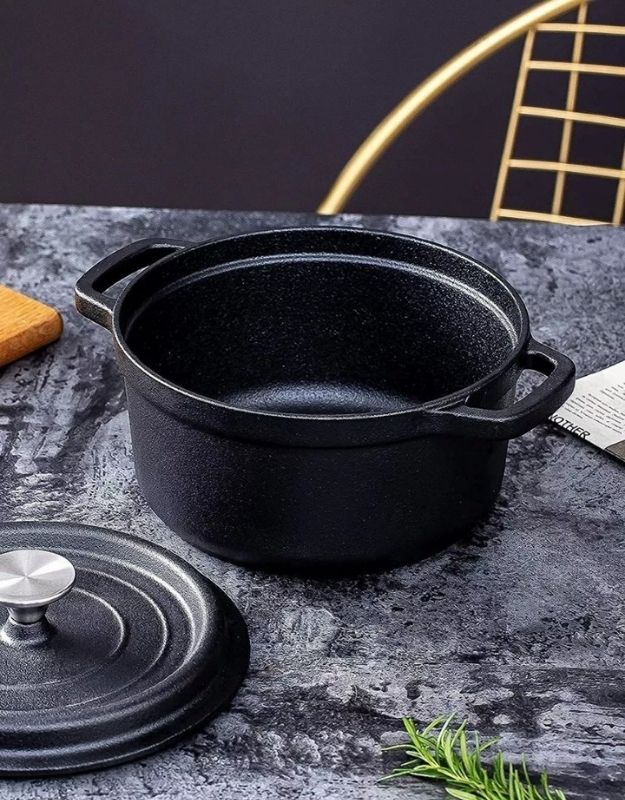 Black cast iron pot on a dark surface with a black background. Black cast iron cooking pot with lid placed beside it on a gray kitchen table.