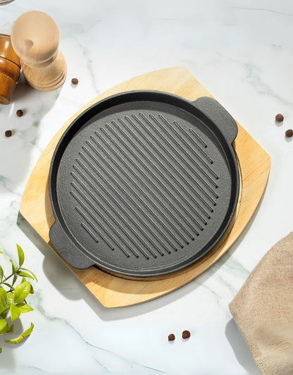 A round, ribbed cast iron griddle pan on a wooden base, with a bamboo stand and various cooking utensils in the background.