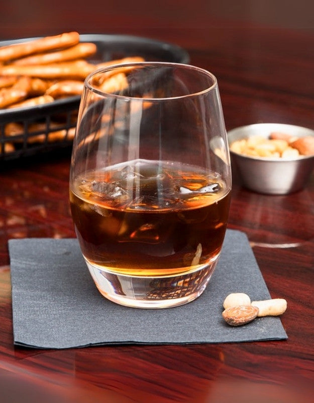Glass of amber liquid on a dark napkin with snacks in the background. Clear round whiskey glass with drink and ice cubes on wooden table.