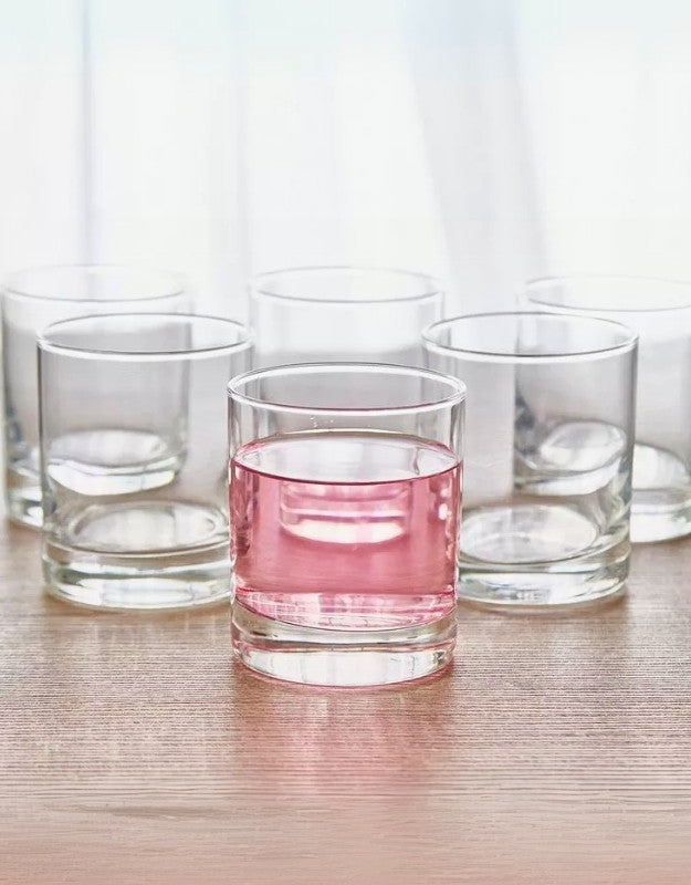 Set of six clear short drinking glasses, one filled with pink beverage on wooden surface.