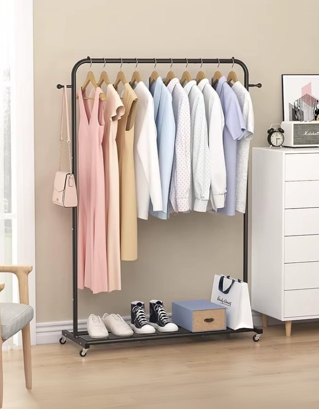A black metal clothing garment rack with clothes hanging on it, placed on wheels and situated in a room corner.