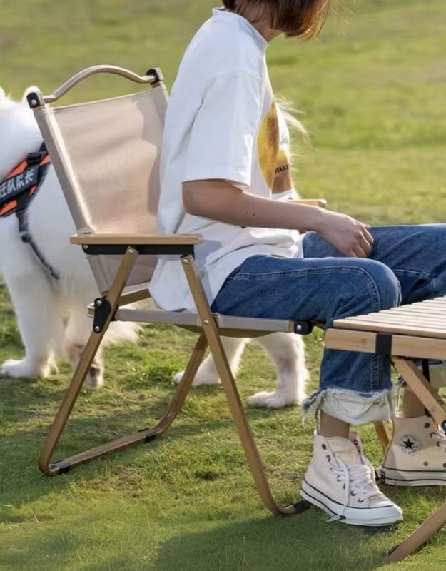 A person is sitting on a foldable camping chair with a light wooden or bamboo frame and beige fabric backrest. They are outdoors on a grassy area beside a small foldable table, with a white dog nearby. The setting suggests a relaxed, casual outdoor environment, ideal for picnics or camping.