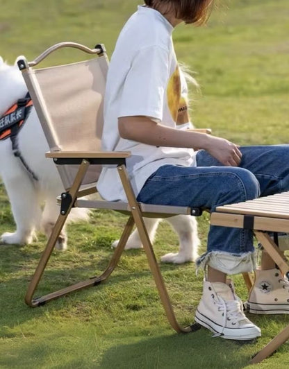 A person is sitting on a foldable camping chair with a light wooden or bamboo frame and beige fabric backrest. They are outdoors on a grassy area beside a small foldable table, with a white dog nearby. The setting suggests a relaxed, casual outdoor environment, ideal for picnics or camping.