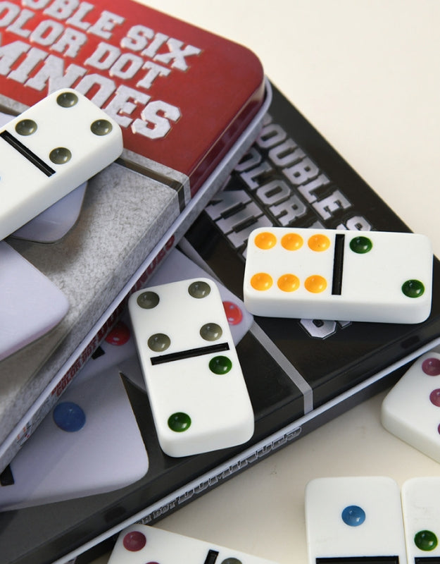 A close-up, angled shot of several domino pieces from the "Double Six Color Dot Dominoes" set, with the open tin box visible in the background