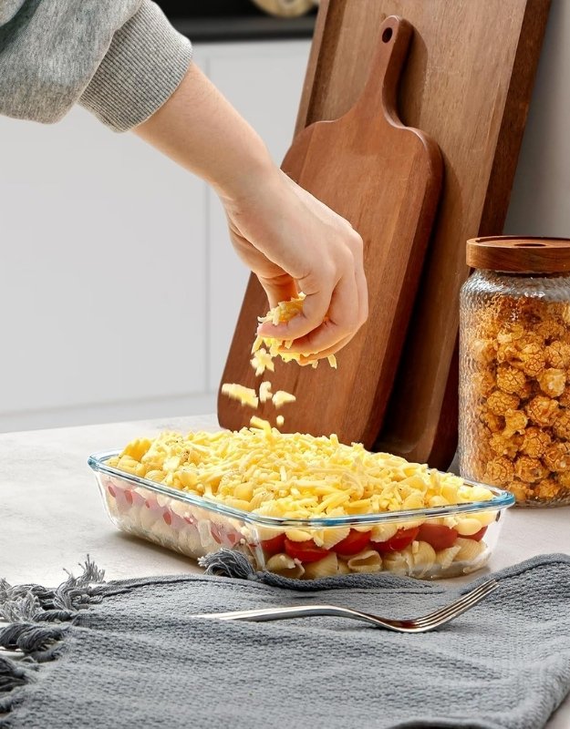 Rectangular tempered glass baking dish being filled with grated cheese for oven cooking