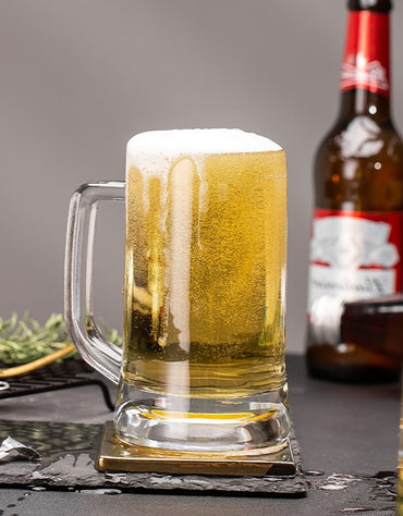 Glass mug filled with beer next to a bottle on a dark surface. Frosted glass beer mug filled with golden lager and foam head.