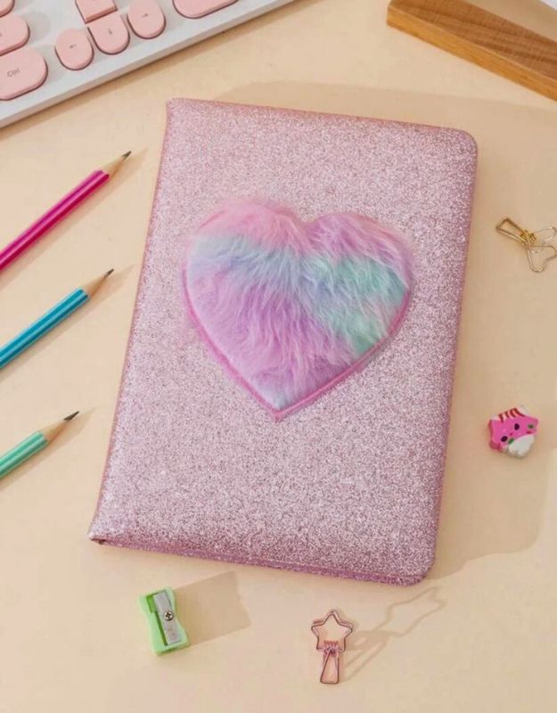 Pink glittery notebook with a heart-shaped fluffy cover on a desk with stationery items.