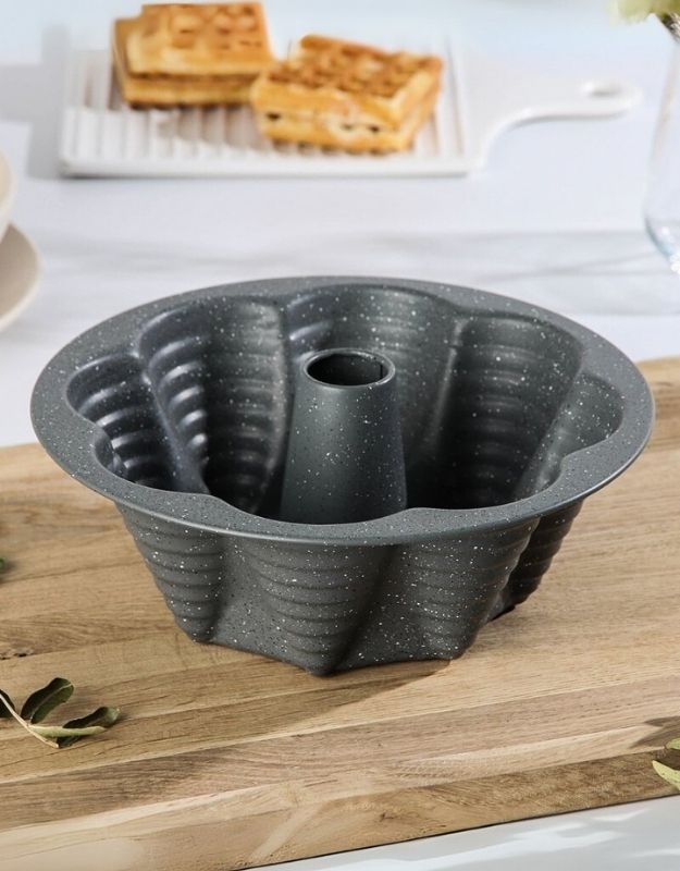 Bundt cake pan on a wooden surface with waffles in the background. Gray non-stick bundt cake pan with fluted design on wooden board.