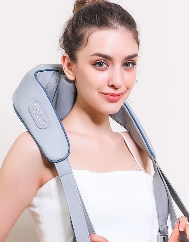 A woman is using a light gray electric neck and shoulder massager. The device has control buttons on one side and straps for easy handling. She looks relaxed and comfortable, wearing a white sleeveless top.