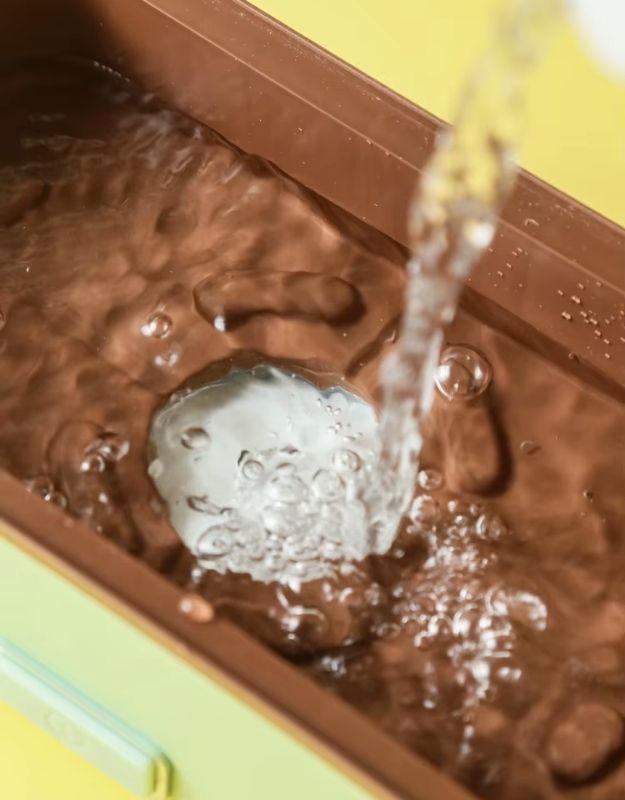 Water being poured into the heating compartment of an electric lunch box.
The water is likely used to create steam, which gently heats the food placed in the stainless steel containers above.