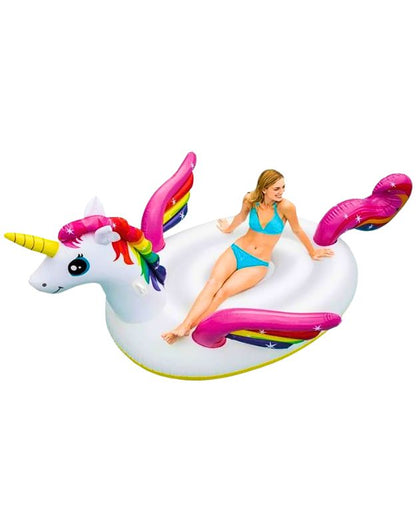 Model posing on oversized white unicorn inflatable with rainbow wings and horn on plain background.
