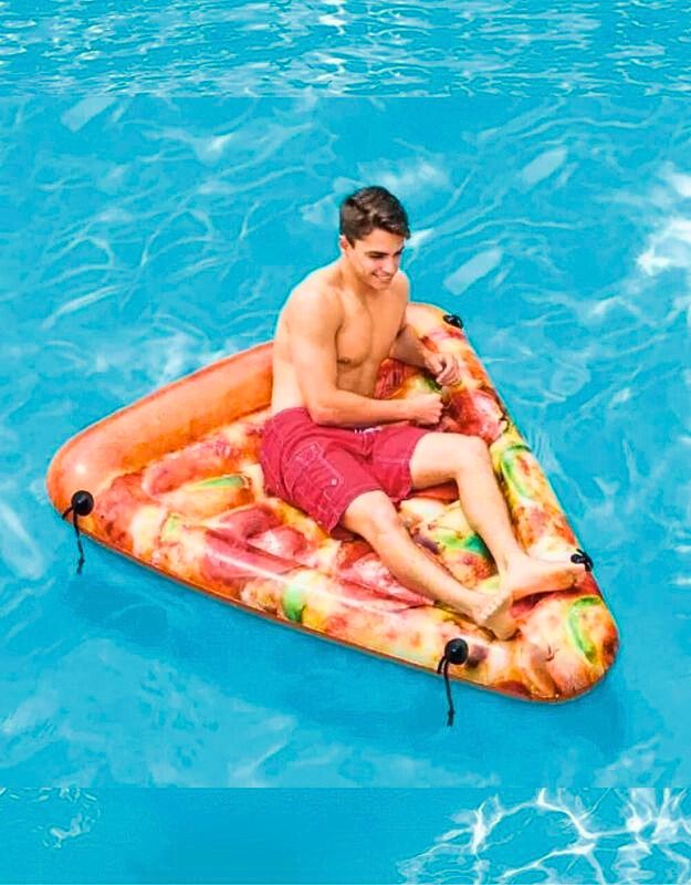Man sitting on an inflatable pizza slice float in a swimming pool, wearing red swim short