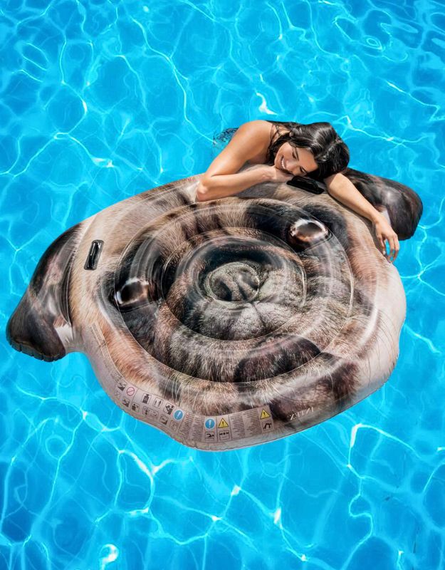 Woman relaxing on an Intex realistic inflatable pool float designed with a pug dog face, floating on clear blue water.