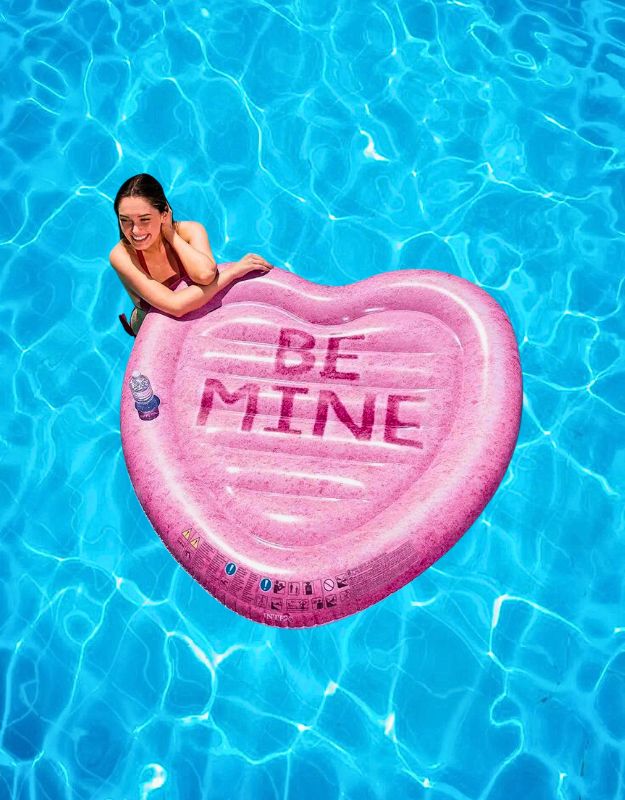 A woman in a purple bikini enjoying the pool while leaning on a large pink heart-shaped inflatable float with the words "BE MINE" written on it.