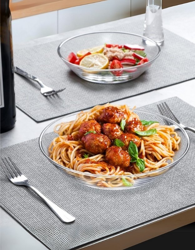 Two glass cocktail plates on dining table, one filled with spaghetti and meatballs, the other with salad and lemon slices.