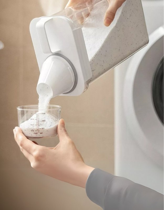 White dispenser pouring liquid into a glass container held by a person. Airtight dispenser container pouring laundry detergent powder into measuring cup.
