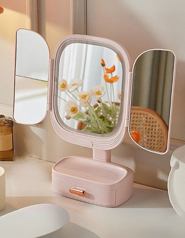 Pink trifold vanity mirror with LED light placed on dressing table with flowers.