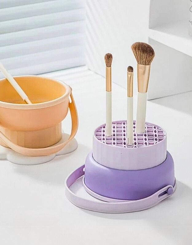 A purple silicone makeup brush cleaner bowl with brushes being cleaned inside it, placed on a white surface next to a yellow bucket.