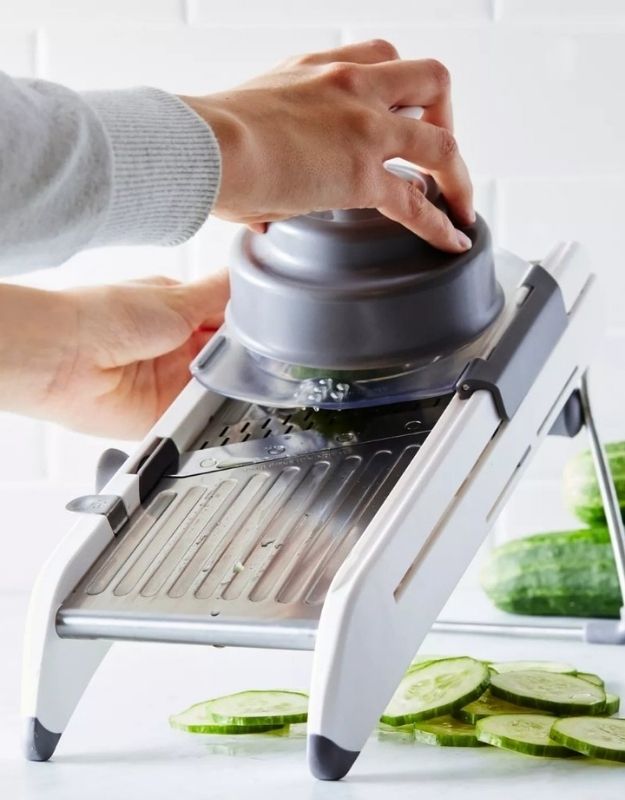 Person using a mandoline slicer to slice cucumbers on a white surface. Mandoline slicer cutting cucumber slices with protective holder.