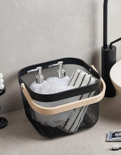 Black mesh storage basket with wooden handle holding bathroom toiletries and towels.
