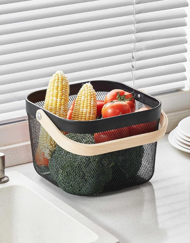 Black mesh storage basket with wooden handle filled with fresh vegetables on a kitchen counter.