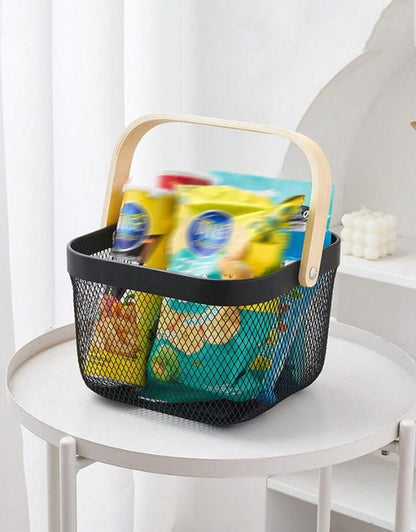 A black metal mesh basket with a wooden handle filled with colorful items, placed on a white table.