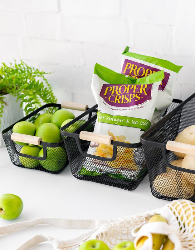 Set of rectangular mesh baskets with wooden handles holding apples, crisps, and potatoes on counter.
