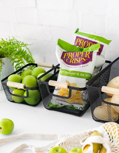 Set of rectangular mesh baskets with wooden handles holding apples, crisps, and potatoes on counter.