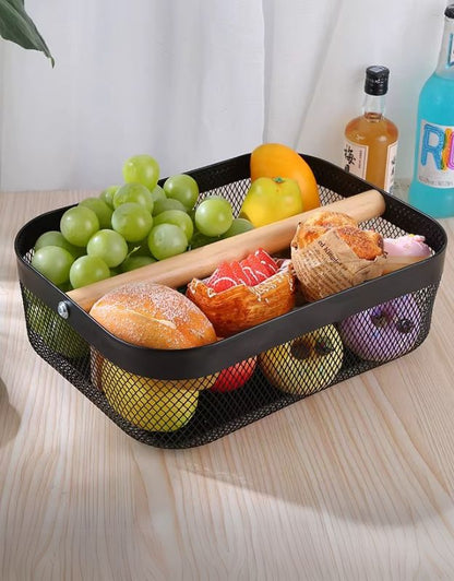 A black metal mesh basket with a wooden handle being used to hold various fruits and snacks inside it, placed on a wooden table.