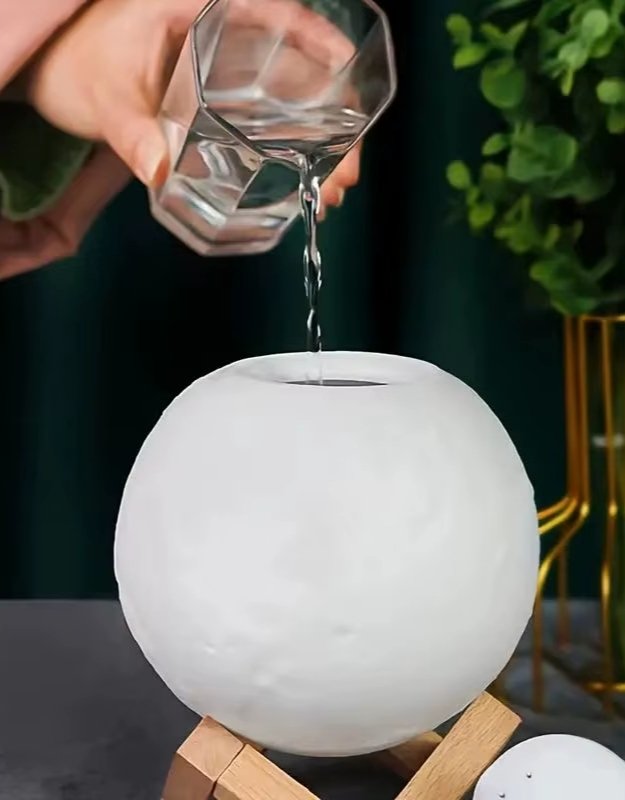 Glass of water being poured into white moon lamp humidifier on wooden stand.