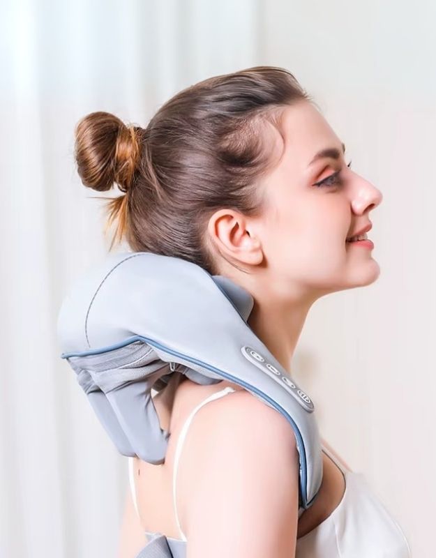 A woman is using a light gray electric neck and shoulder massager. The massager wraps around her neck and shoulders, with control buttons visible on one side